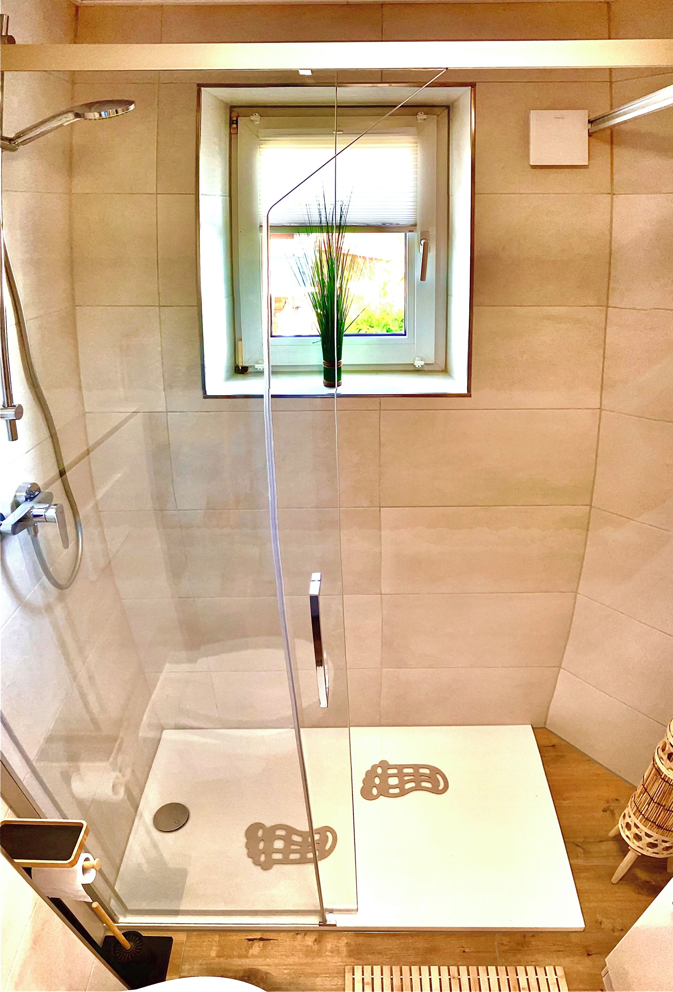 Une douche fermée par des parois de verre avec une fenêtre, une petite table agrémentée d’un objet décoratif et un plancher en bois.