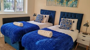 A hotel room with two beds, each with a blue bedspread and white linens, a large abstract painting on the wall, and a window with a view of buildings. - Newly refurbished One Bedroom House With Front Garden (London)