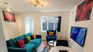 A modern living room with a teal sofa, a blue armchair, a round coffee table, and a flat-screen TV. - Newly refurbished One Bedroom House With Front Garden (London)