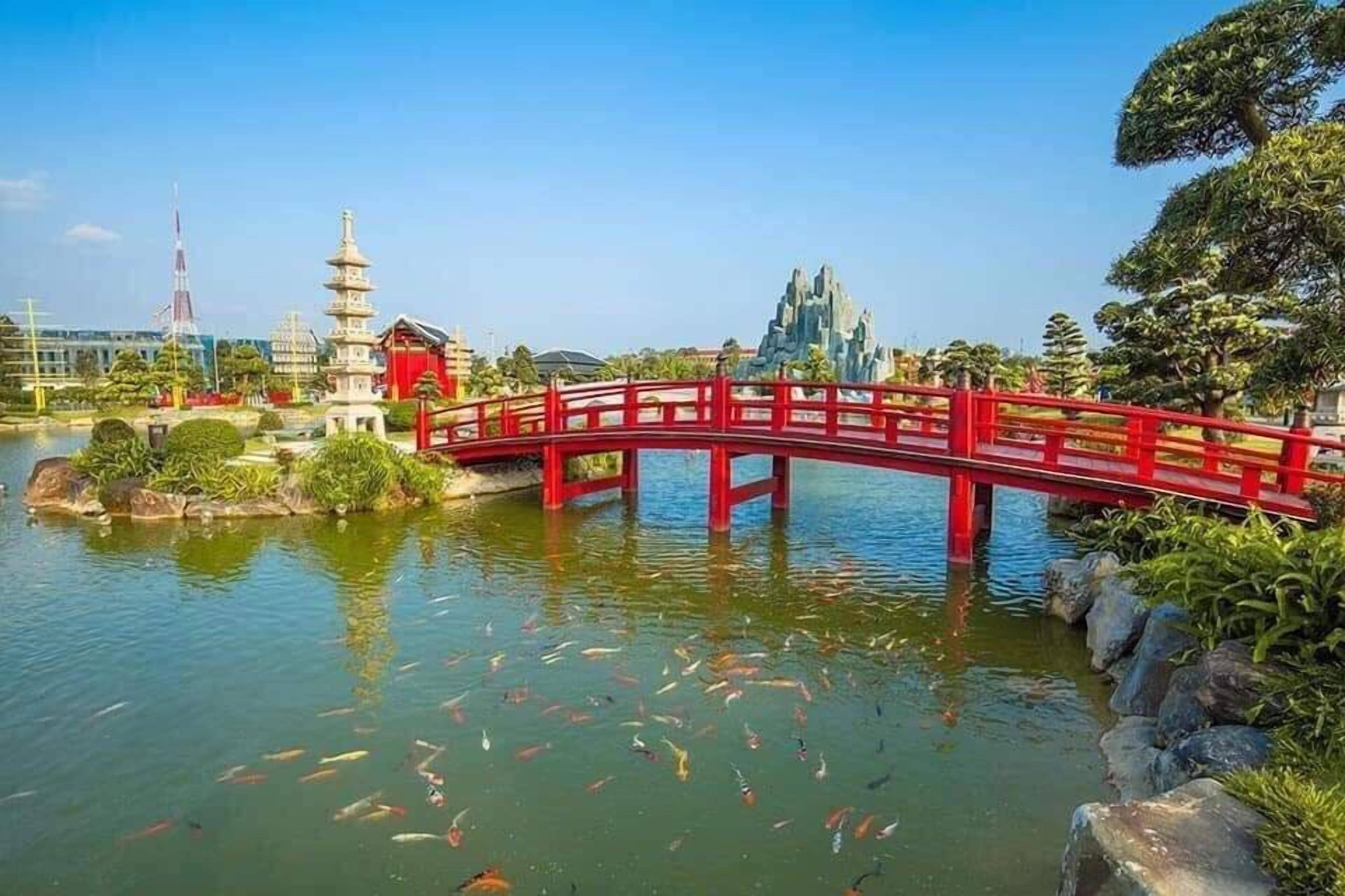 Een rode brug over een vijver met koi karpers, omgeven door begroeiing en een pagode-achtige constructie.