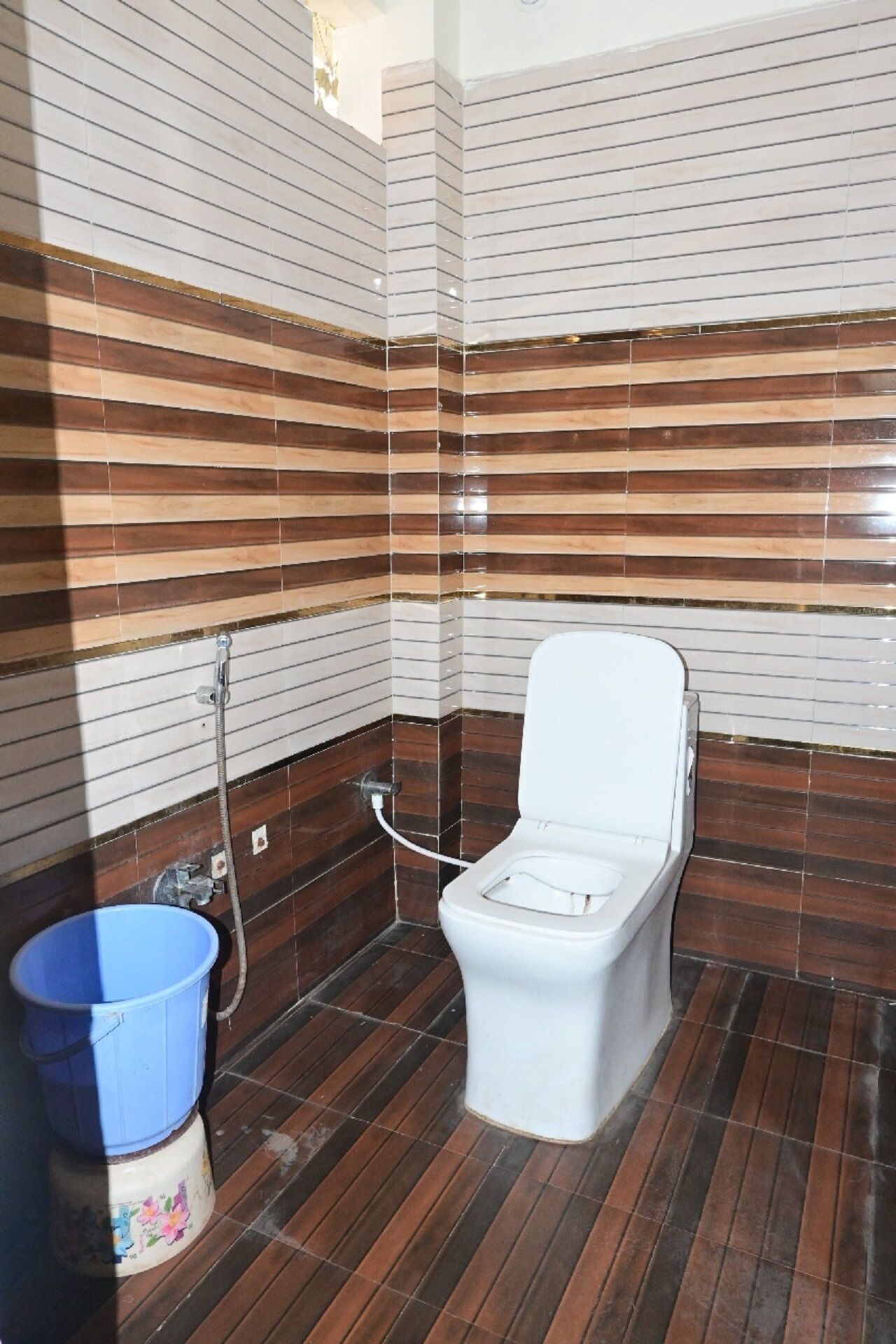 Ein Badezimmer mit einer weißen Toilette, einem blauen Eimer und Wandfliesen mit Streifen.