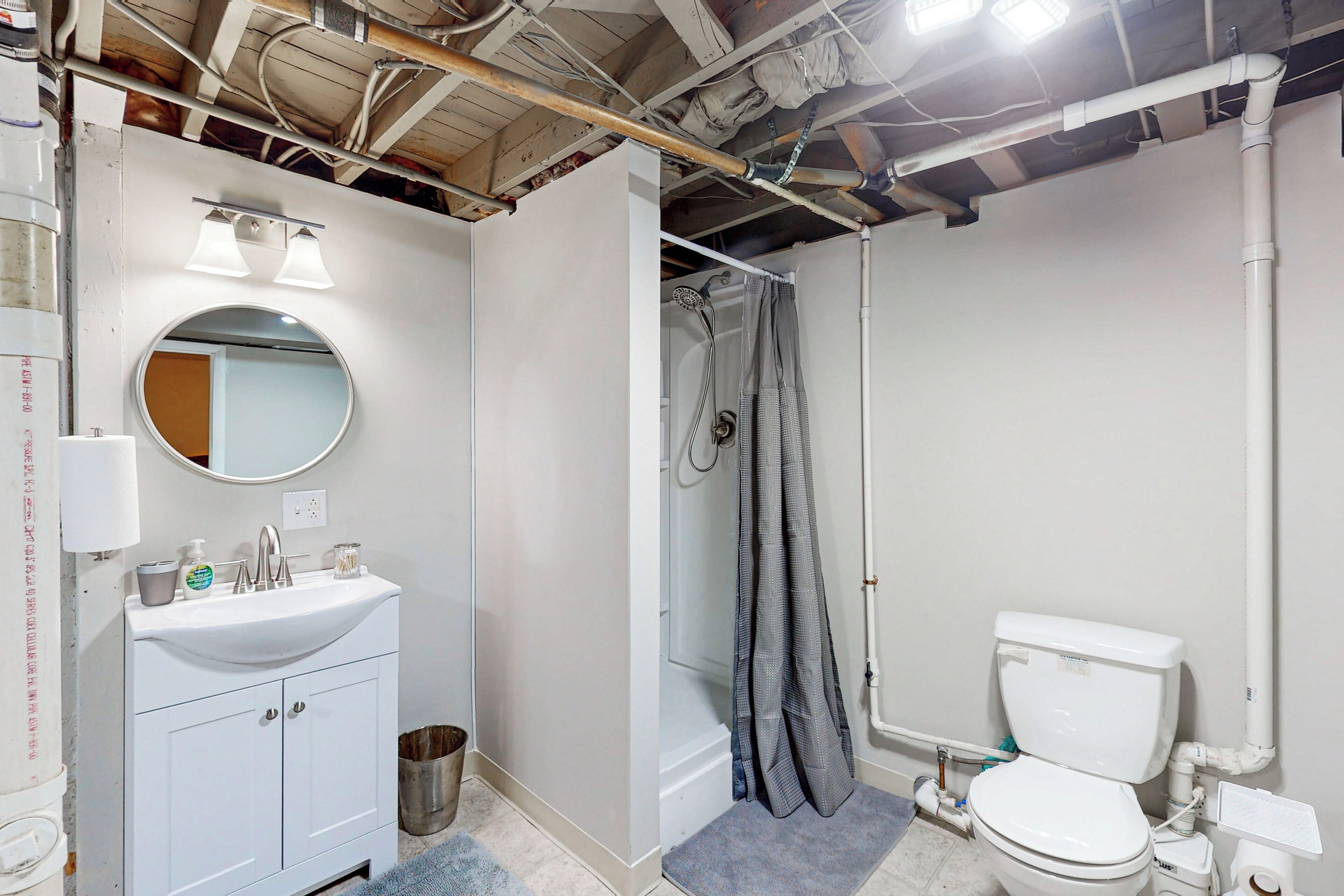 Une salle de bain avec un lavabo blanc, un miroir et des toilettes. On y trouve un rideau de douche et une poubelle.