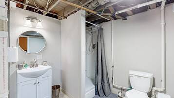 Une salle de bain avec un lavabo blanc, un miroir et des toilettes. On y trouve un rideau de douche et une poubelle.