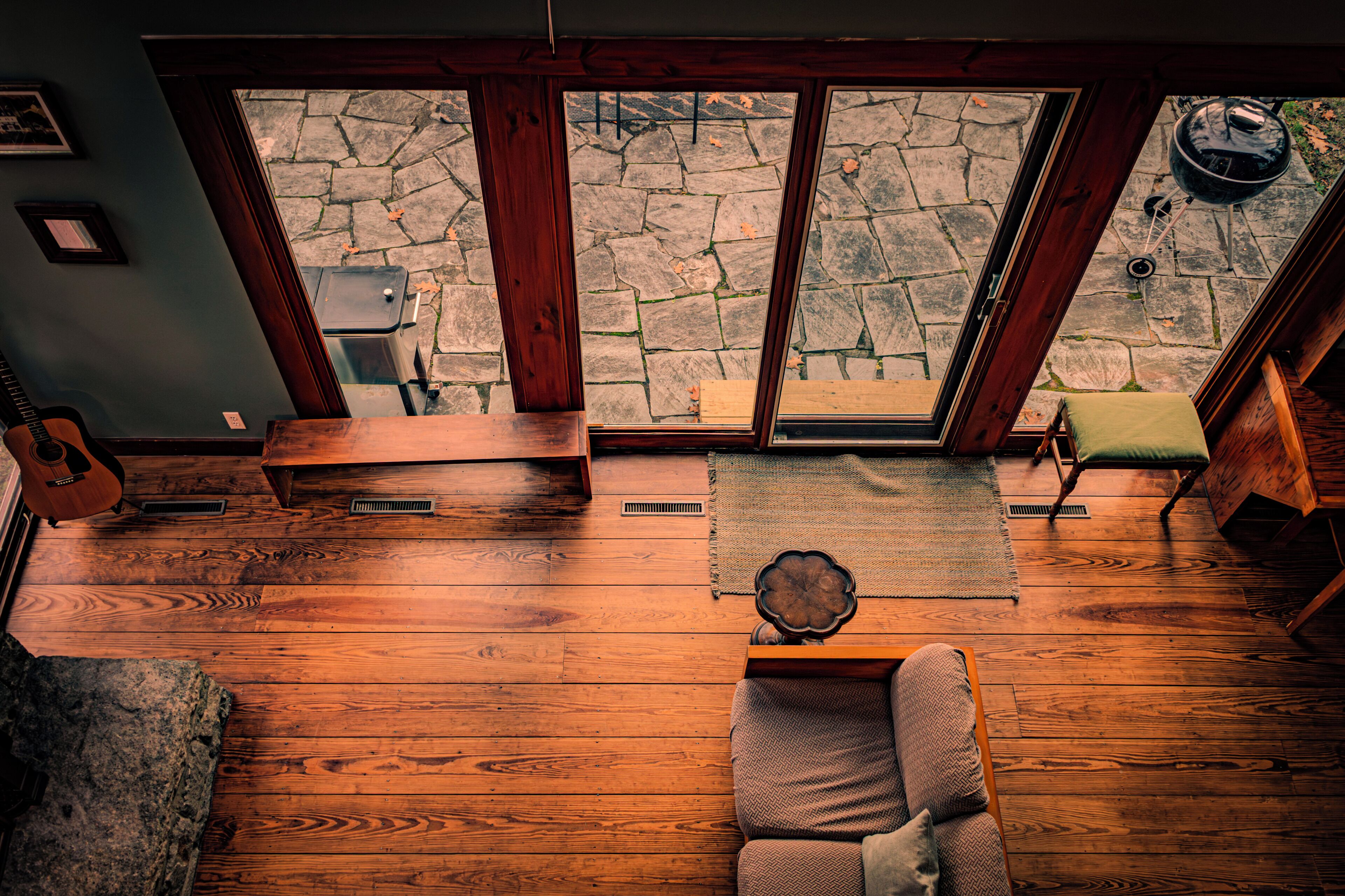 Una habitación acogedora con suelo de madera, un sofá verde con cojines, una mesa de centro de madera y un patio con baldosas de piedra visible a través de amplios ventanales.