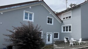 Un bâtiment à deux étages, doté d’une façade grise, de fenêtres à cadre blanc, d’une porte ornée d’un vitrail en forme de losange et d’un petit jardin avec des chaises blanches et un banc en pierre.