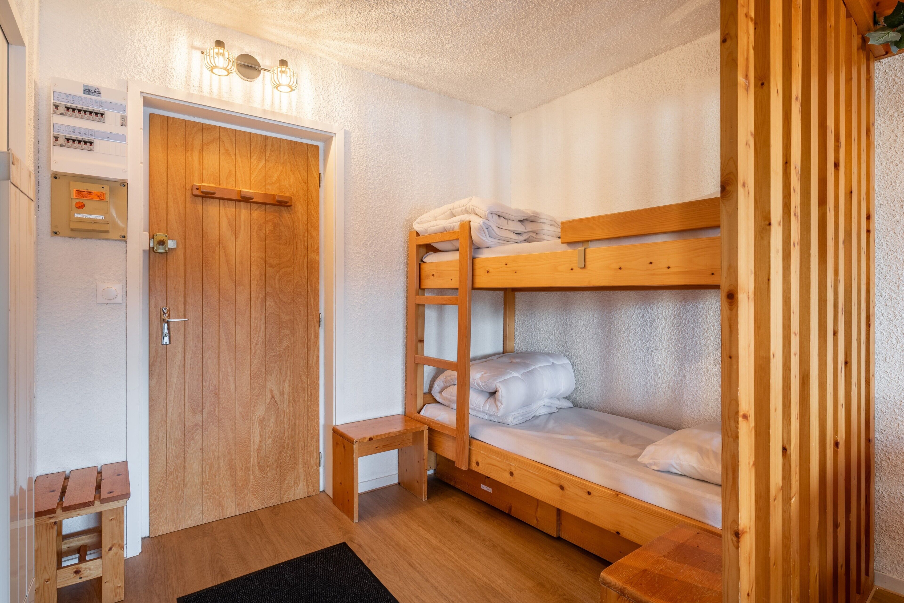 Een slaapkamer met stapelbed, een houten deur, een klein houten bankje en een lichtschakelaar aan de muur.