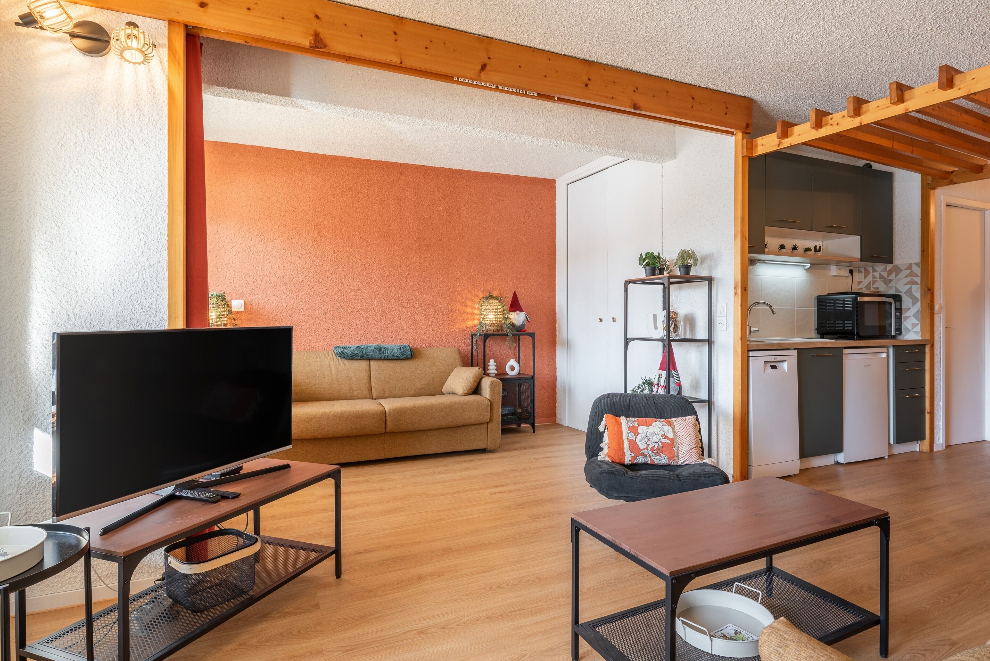 A living room with a TV, sofa, and coffee table, a kitchen with appliances, and a wooden ceiling.