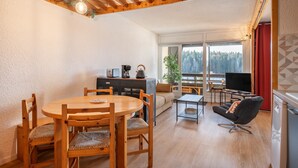 Une table à manger en bois avec des chaises, un canapé, un téléviseur et un balcon avec vue sur les arbres.