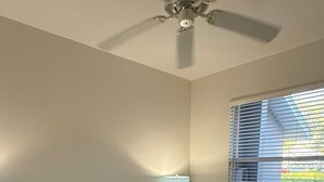 Ein Schlafzimmer mit einem Bett, einem Deckenventilator, einem Nachttisch mit Lampe und einem Fenster mit Jalousien.
