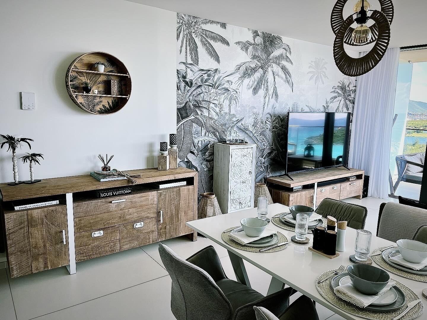 A modern dining area with a white table set for four, a wooden cabinet with a Louis Vuitton sign, a large tropical mural, and a TV on a wooden stand.