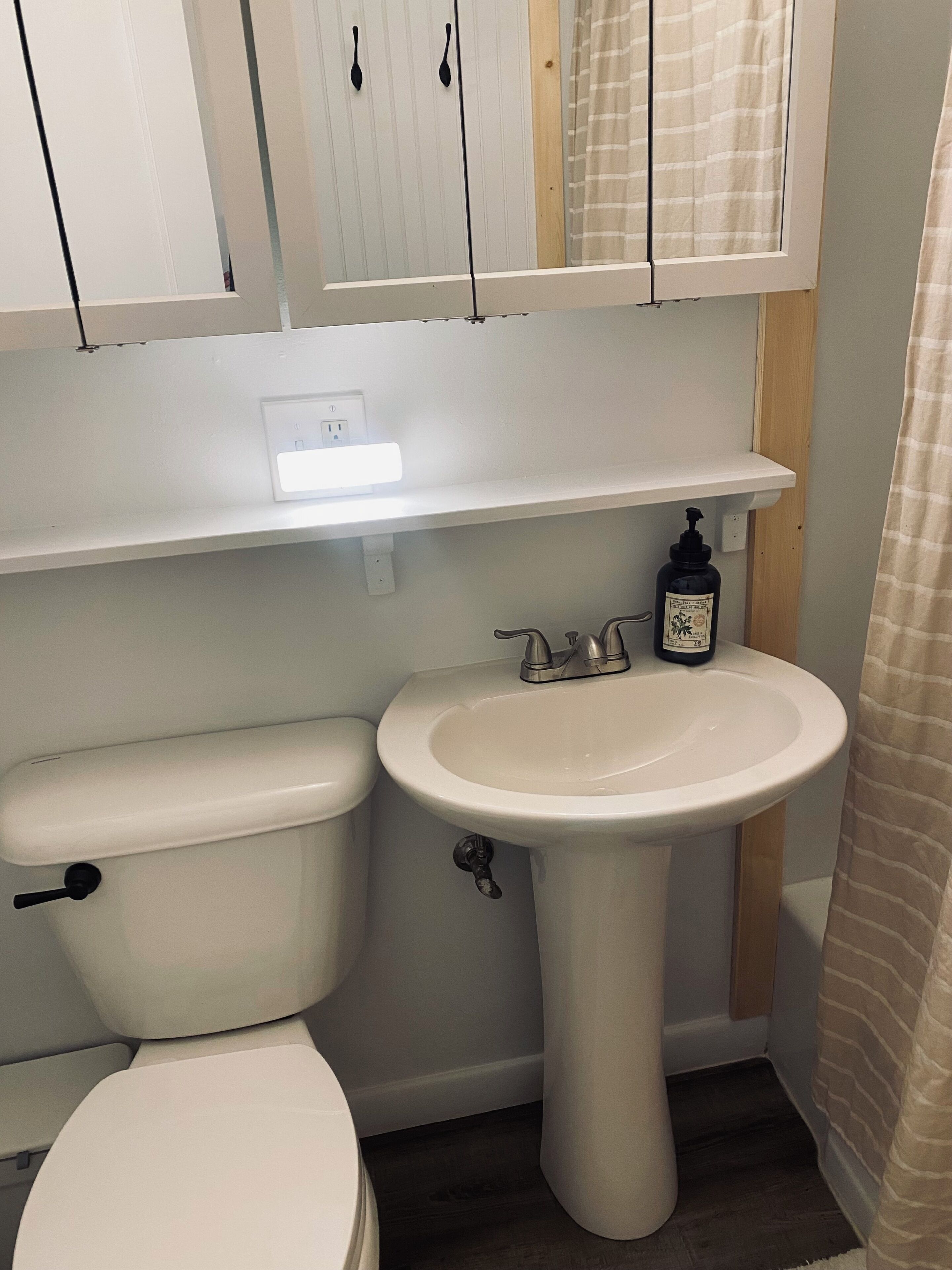 Un baño con inodoro blanco, lavamanos sobre pedestal y repisa arriba del lavamanos.