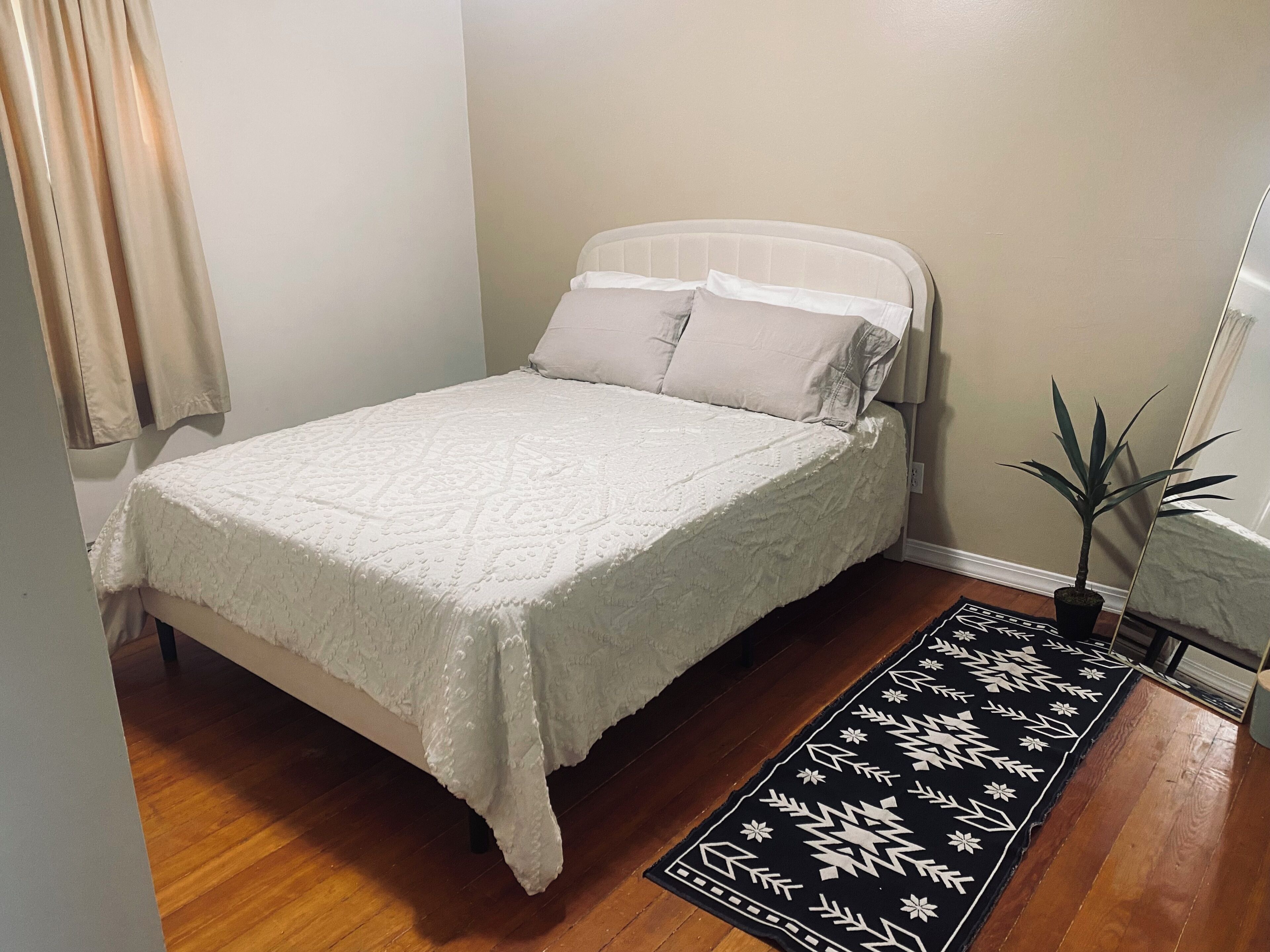 Una cama bien hecha con una colcha blanca y almohadas, suelo de madera, alfombra estampada en blanco y negro y una planta en maceta.