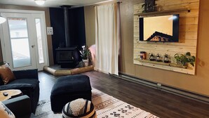 A cozy living room with a fireplace, a TV mounted on a wooden wall, and a comfortable seating area with a rug and cushions. - Charming 2-BR, no cleaning fees, cozy fireplace (Plains)