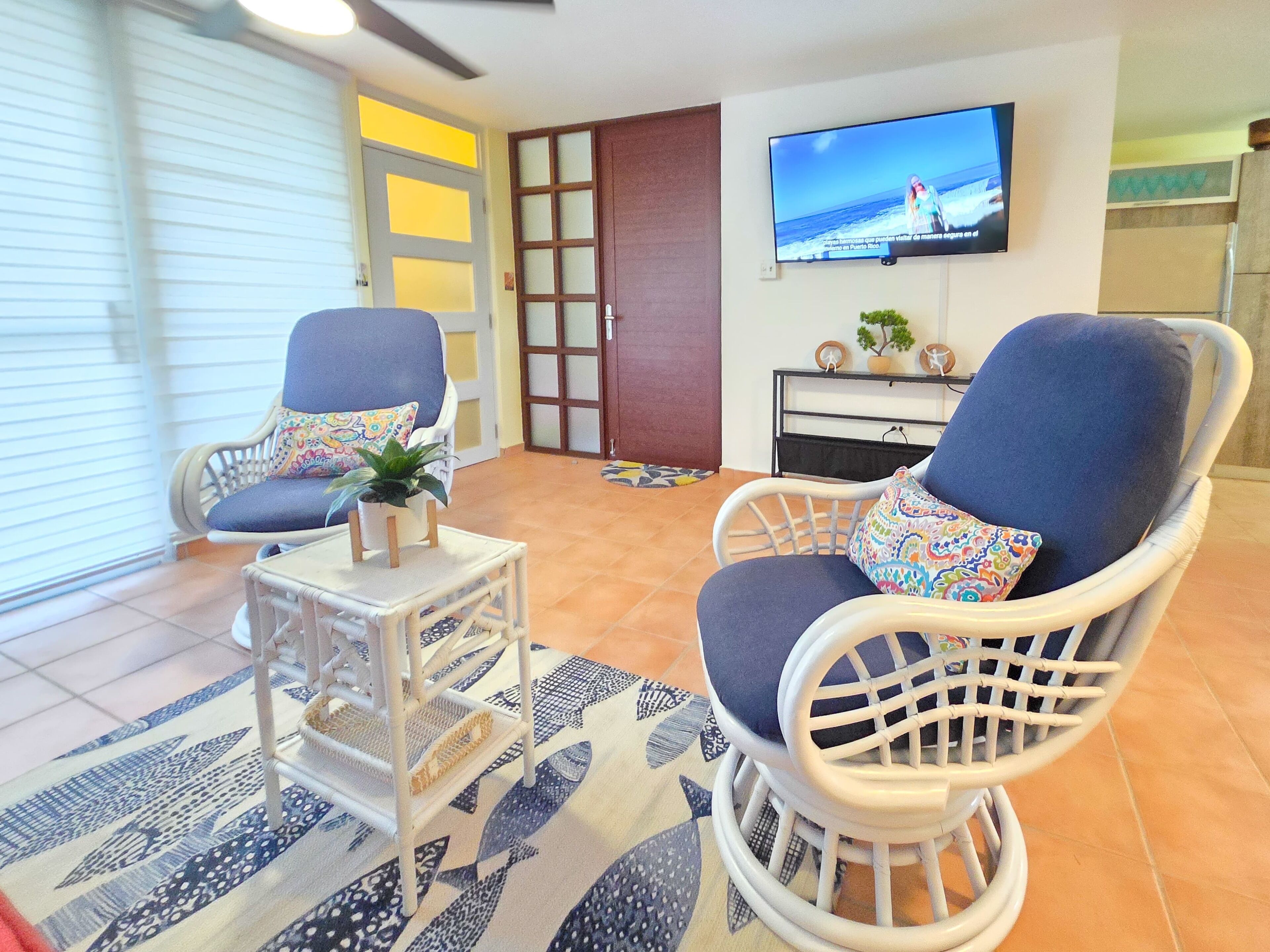 A living room with two wicker chairs, a small table with a plant, and a television on the wall.