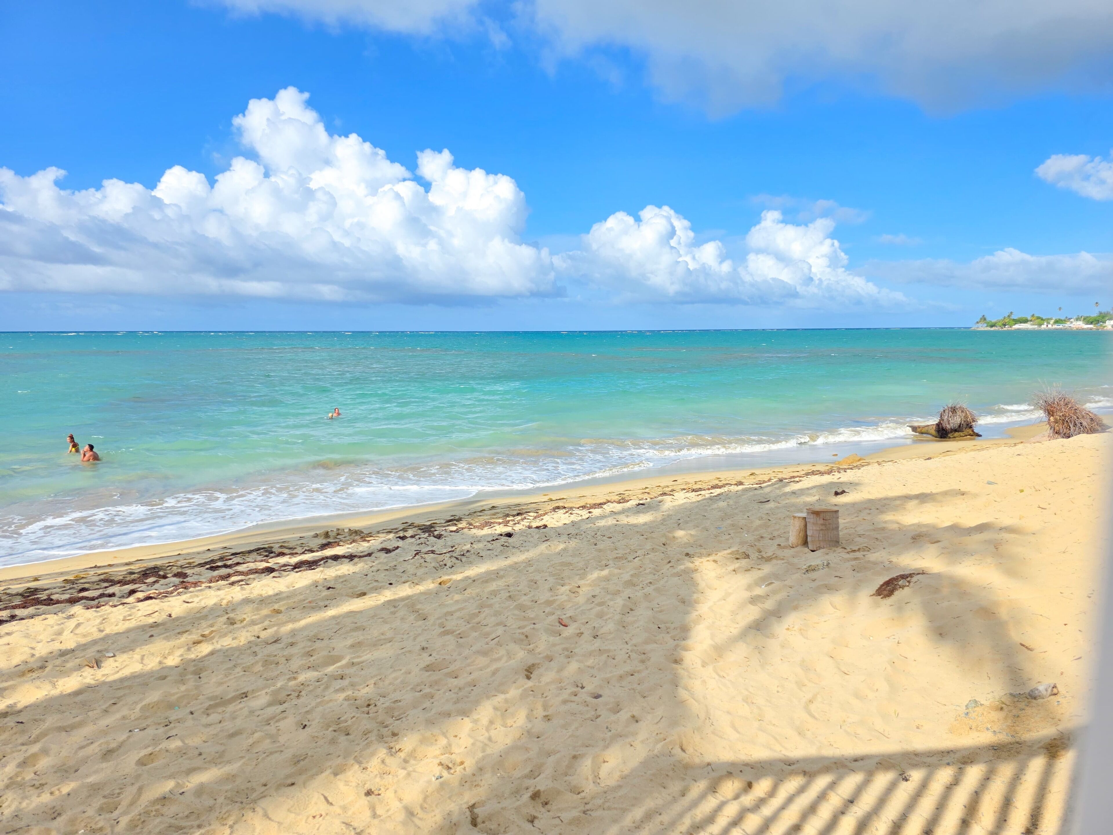 A beach with clear turquoise water, a sandy shore, and a few people swimming.