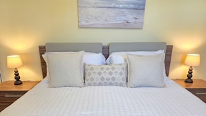 Cama bem arrumada com colcha branca, quatro travesseiros e duas luminárias de cabeceira em mesas de cabeceira de madeira.