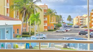 A balcony with a view of a swimming pool, palm trees, and a street with parked cars.