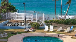 A swimming pool with a curved edge, surrounded by a white fence, with a view of the ocean and palm trees in the background.