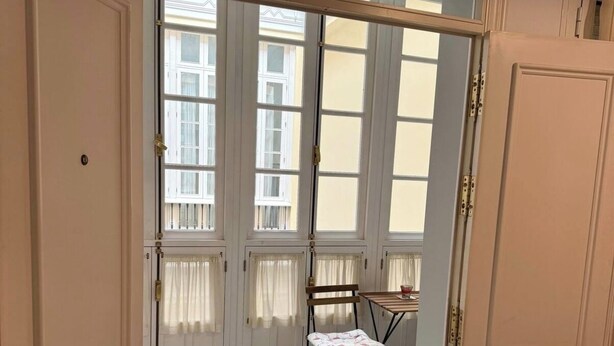 Une pièce dotée d’une grande porte-fenêtre coulissante qui donne sur un balcon offrant une vue sur les immeubles. On y trouve une petite table et deux chaises.