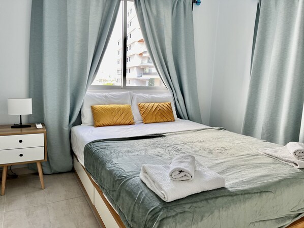 Un lit bien fait, avec une couette grise, deux oreillers de couleur moutarde et une serviette blanche pliée sur le dessus. On aperçoit une table de chevet avec une lampe et un petit appareil. La pièce est dotée de rideaux gris clair et offre une vue sur les bâtiments à l’extérieur.