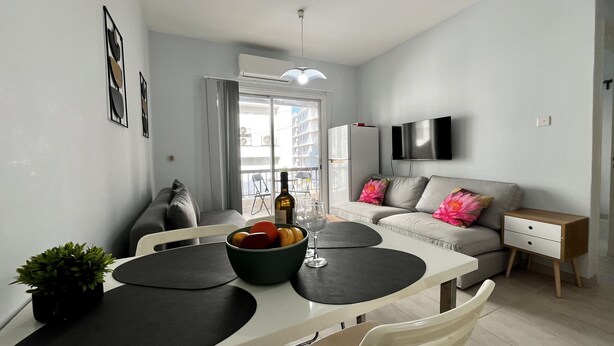A modern living room with a dining area, a grey sofa with floral cushions, a flat-screen TV, and a small table with a bottle of wine and fruit. - 114 LCA - Stay Mckenzie Mile (Larnaca)