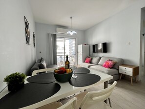 A modern living room with a dining area, a grey sofa with floral cushions, a flat-screen TV, and a small table with a bottle of wine and fruit. - 114 LCA - Stay Mckenzie Mile (Larnaca)