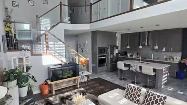 Amplia sala de estar con un sofá blanco, una mesa de centro y un televisor. Se observa una zona de cocina con una barra y una escalera que sube a un nivel superior.