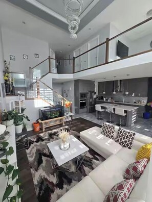 A spacious living area with a white sofa, a coffee table, and a TV. There is a kitchen area with a bar and a staircase leading to an upper level. - Spacious Edmonton Family Rental (Edmonton)