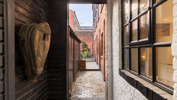 A narrow alleyway with a brick pathway leading to a courtyard, flanked by wooden walls and a white brick building with large windows. - Enamel Works - The Bellows (West Midlands)