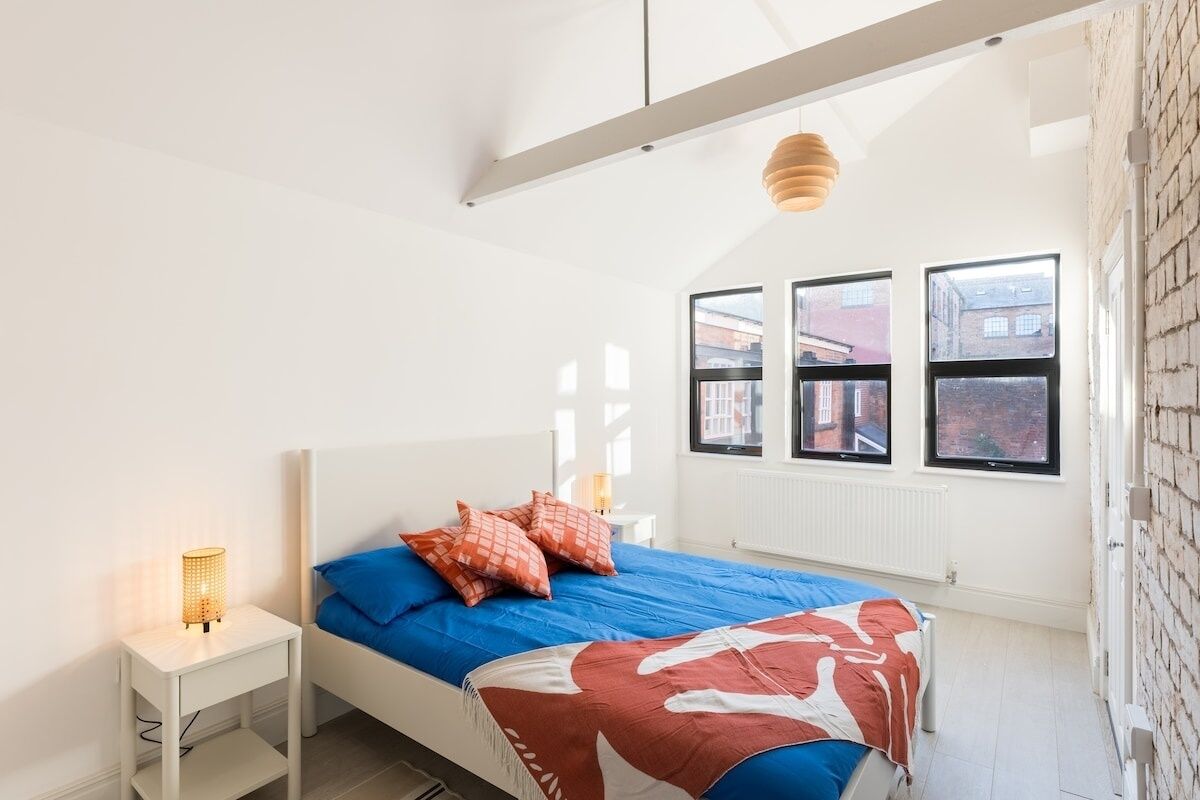 Ein Schlafzimmer mit einem Bett, einem Nachttisch, einer Lampe und einem Fenster mit Blick auf Gebäude.