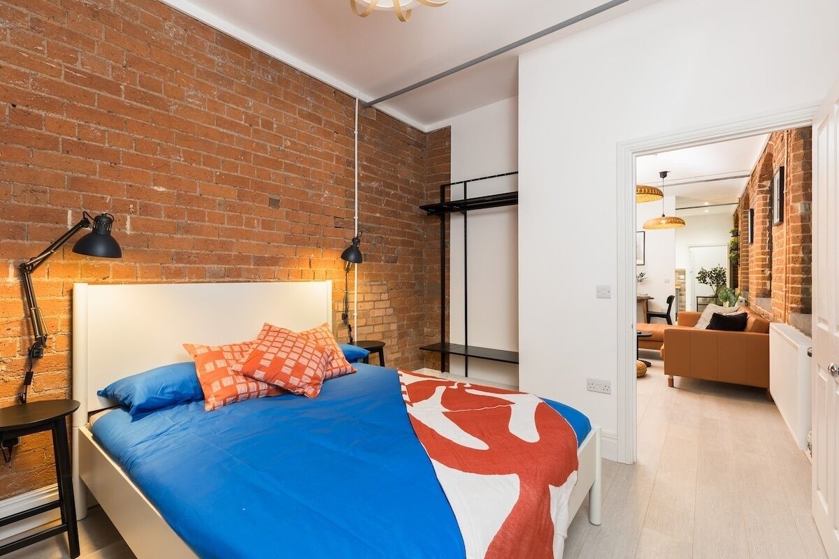 A modern bedroom with a brick wall, a large bed with blue and orange bedding, and a view into the living area.