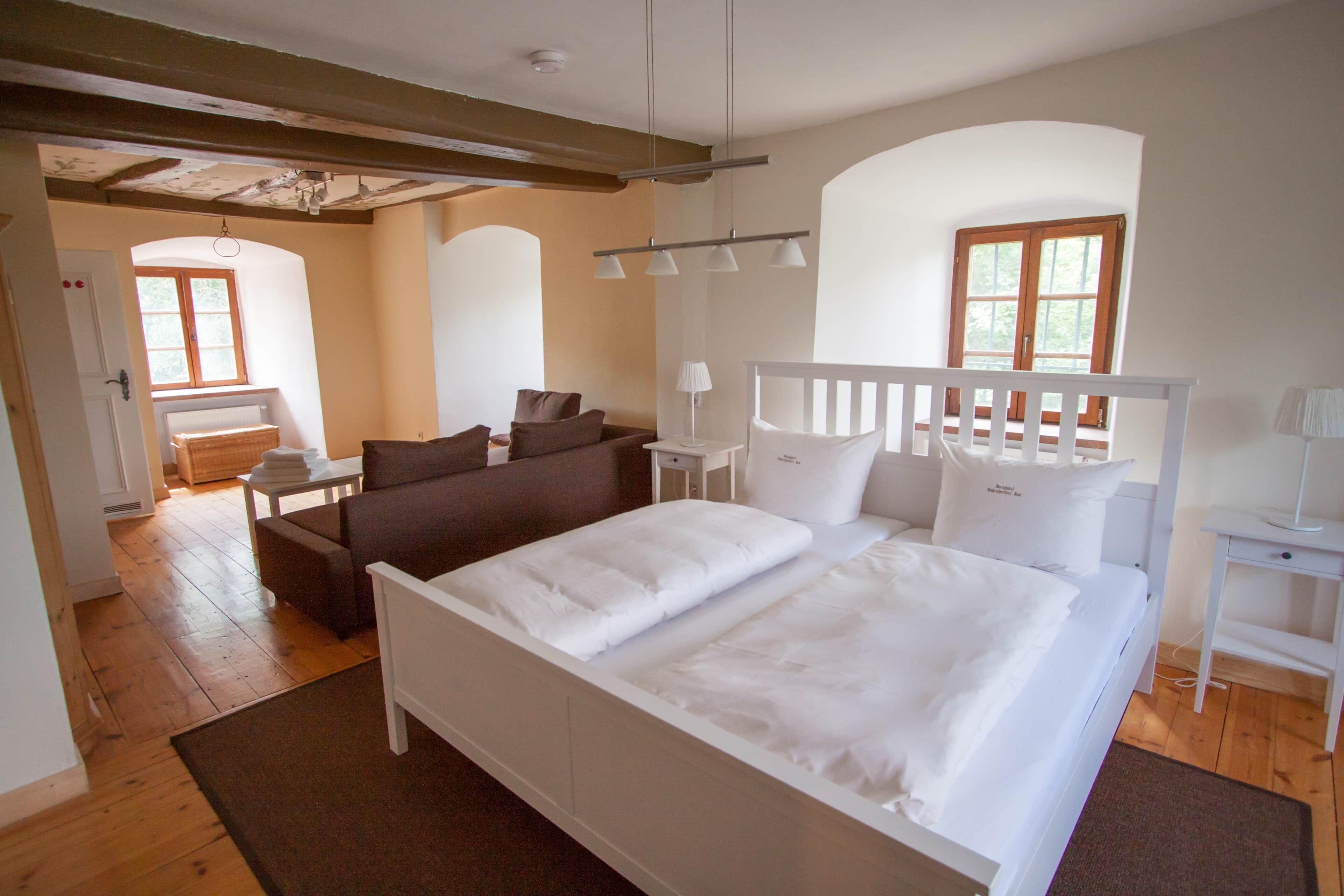Ein Schlafzimmer mit einem großen Bett, einer Couch, einem Schreibtisch und einem Stuhl. Fenster und eine Tür sind zu sehen.