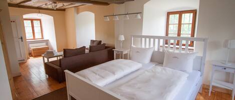 Ein Schlafzimmer mit einem großen Bett, einer Couch, einem Schreibtisch und einem Stuhl. Fenster und eine Tür sind zu sehen.