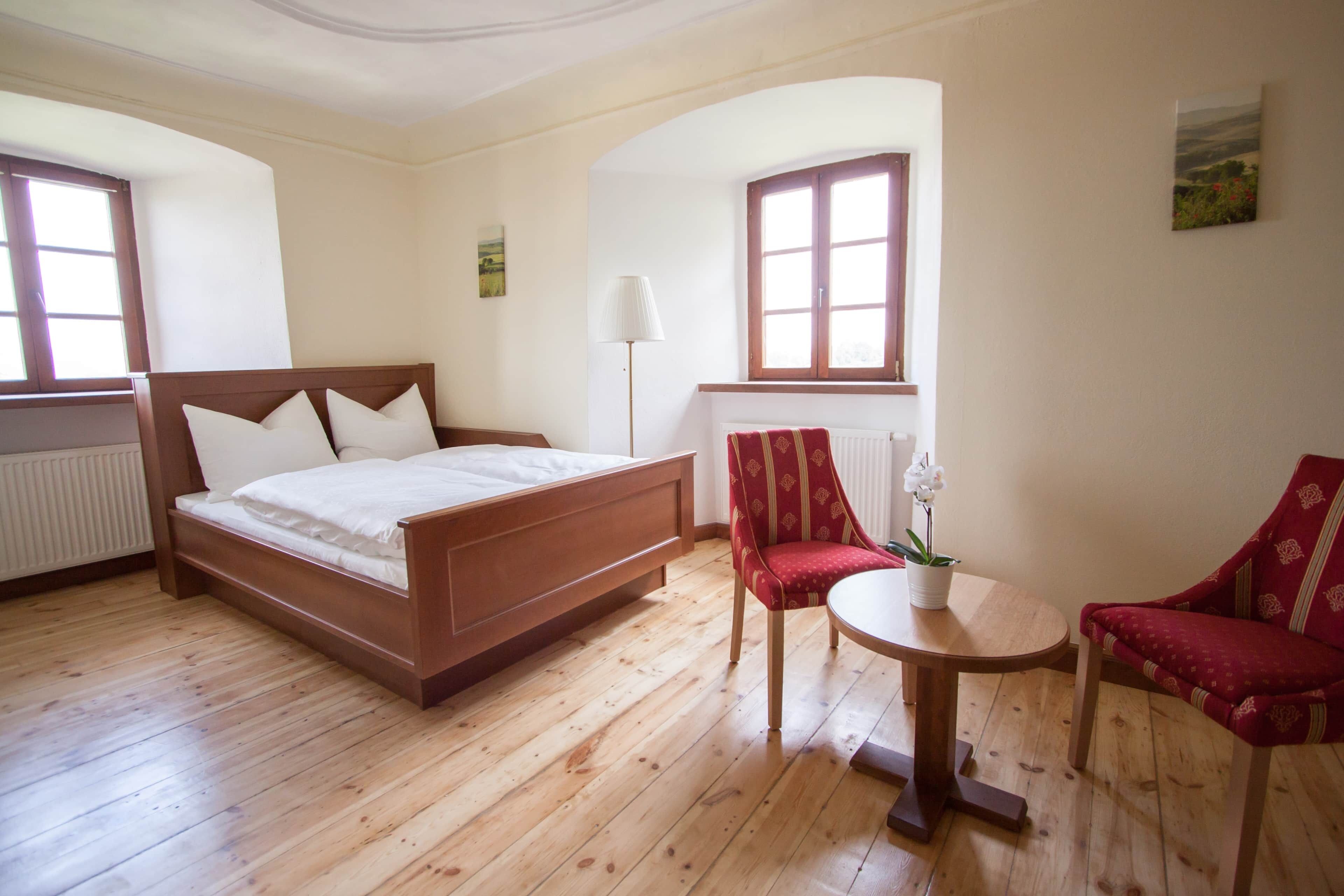 A bedroom with a wooden bed, a red chair, a small table with a potted plant, and a window with a view of the outdoors.