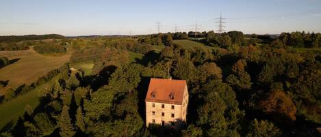 Ein Steinhaus mit rotem Ziegeldach, umgeben von dichtem Wald, mit Stromleitungen und blauem Himmel im Hintergrund.