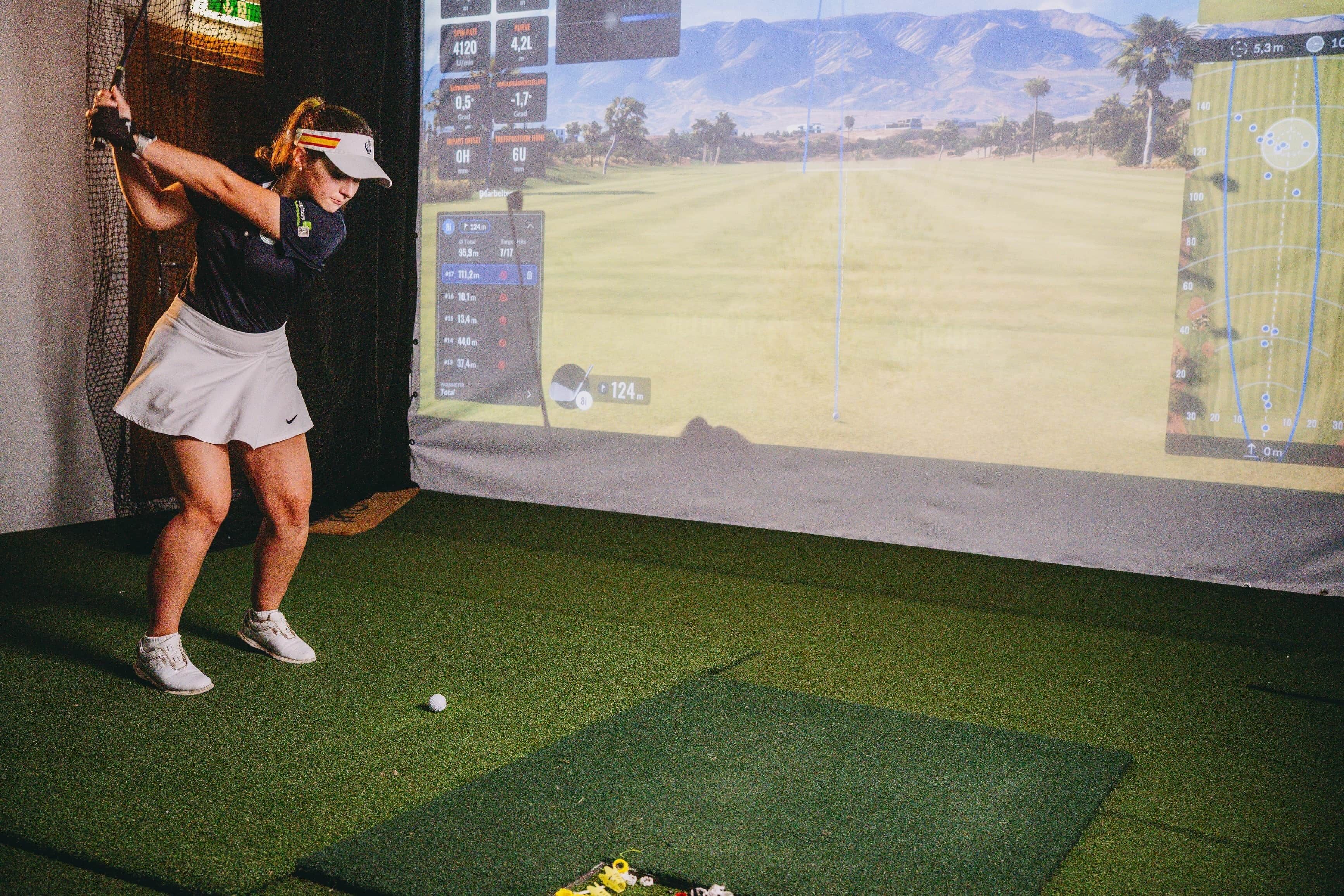 A person practicing golf swings indoors with a virtual golf course displayed on a screen.
