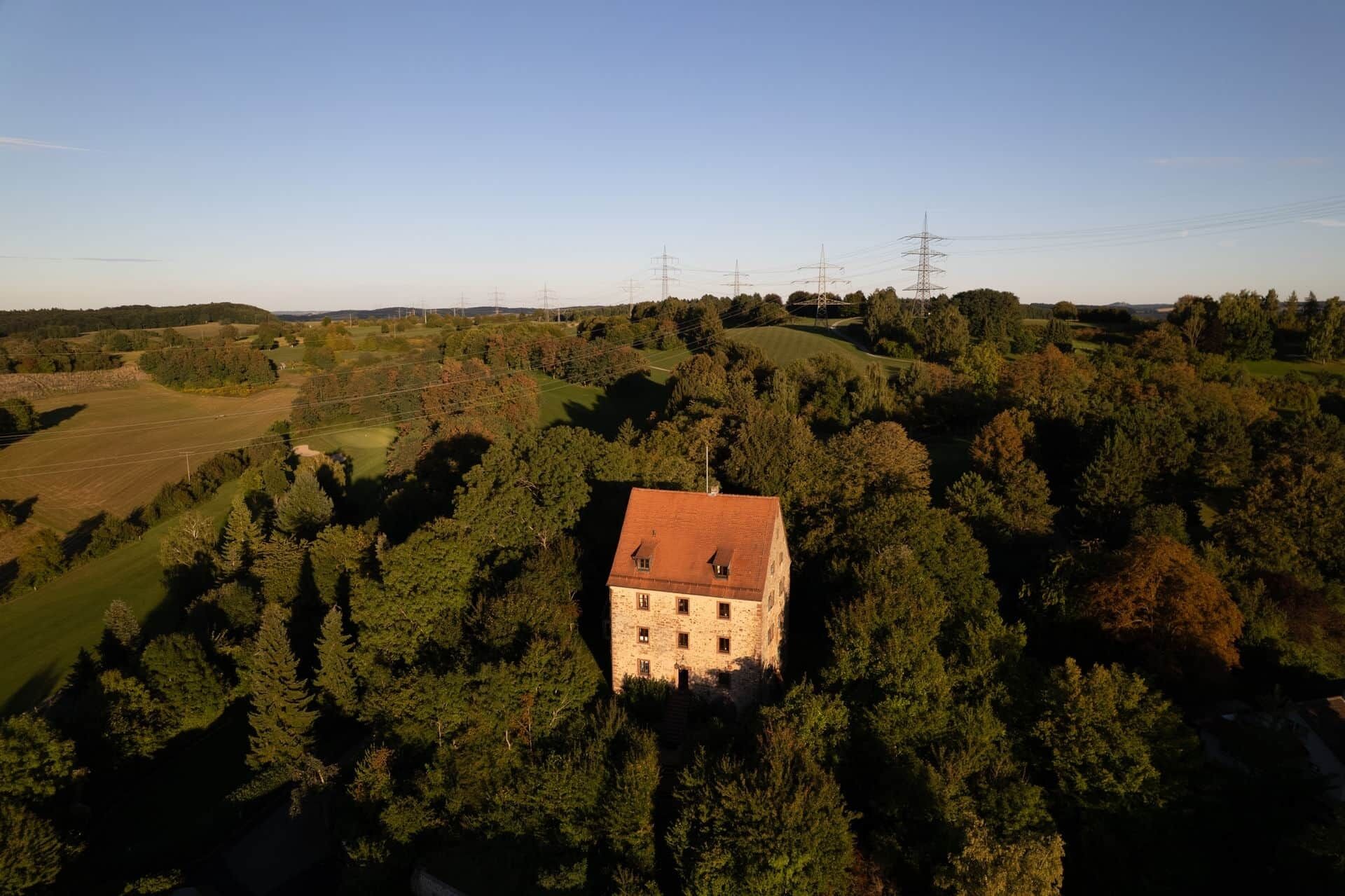 A stone building with a red-tiled roof surrounded by dense forest, with power lines and a clear sky in the background.
