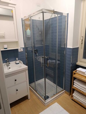 A modern bathroom with a glass shower enclosure, a white sink with a soap dispenser, and a wooden shelf with towels. - Ricc House, Rome (Roma)