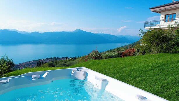 Uma banheira de hidromassagem com vista para um lago e montanhas, cercada por vegetação e uma casa.