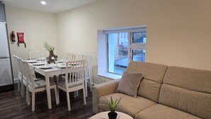 Una sala de estar con un sofá color beige, una mesa redonda de centro con una planta, y un comedor con sillas blancas y una mesa preparada para cuatro.