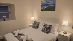 Un dormitorio con una cama, dos lámparas de noche, un ventanal con persianas, una planta en maceta y un cuadro en la pared.