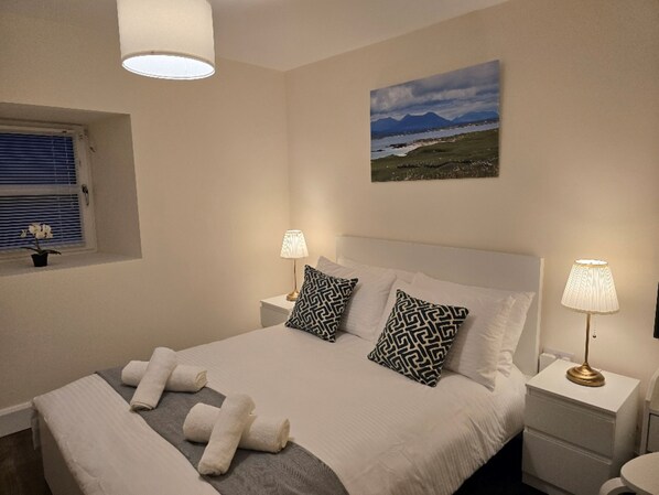 A bedroom with a bed, two bedside lamps, a window with blinds, a potted plant, and a painting on the wall.