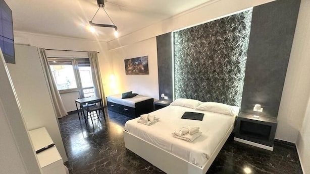 A modern hotel room with a large bed, a desk, a chair, a nightstand, and a decorative wall panel. - Gregory Lem collection (Rome)