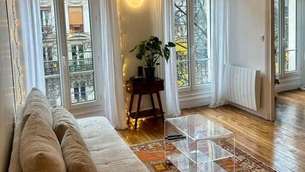 Ein Wohnzimmer mit einem beigefarbenen Sofa, einem Glastisch und einem gemusterten Teppich. Große Fenster mit Vorhängen, ein Blumentopf und eine Wandleuchte sind ebenfalls vorhanden.
