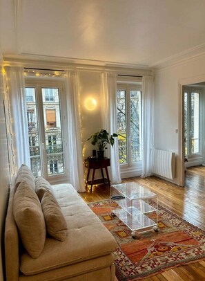 A living room with a beige sofa, a glass coffee table, and a patterned rug. There are large windows with curtains, a potted plant, and a wall-mounted light. - Cosy appartement Montmartre (Paris)