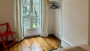 Ein Zimmer mit Holzboden, einem Bett mit beigefarbener Bettwäsche, einem roten Sessel, einem Fenster mit Vorhängen und einer hängenden Lampenleuchte.