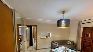 Un coin repas avec une table recouverte d’une nappe à motifs floraux, un canapé avec des coussins, un luminaire suspendu et une petite étagère fixée au mur, sur laquelle sont posés divers objets.