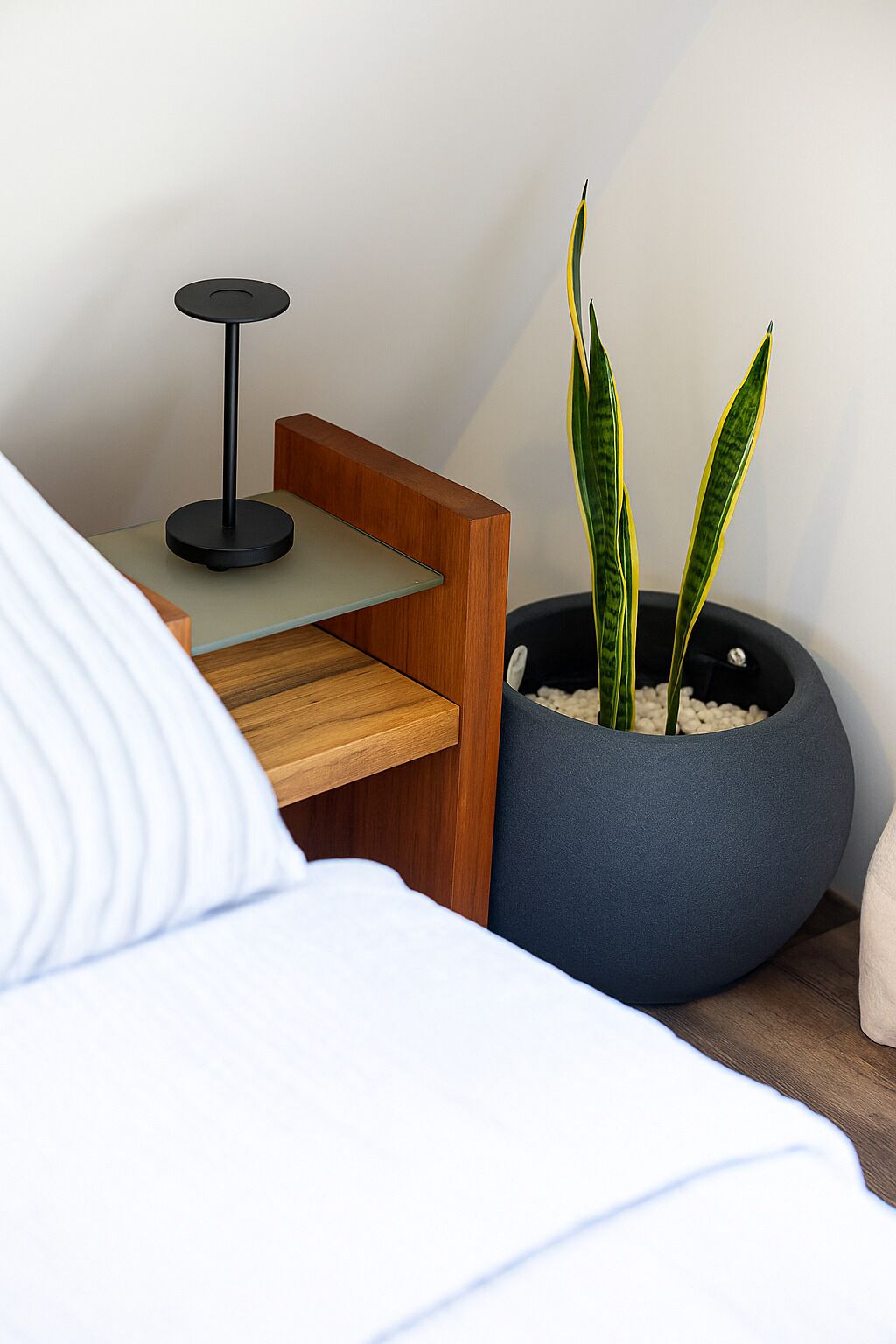 A modern bedroom with a wooden bedside table, a round blue planter with two green plants, and a black floor lamp.