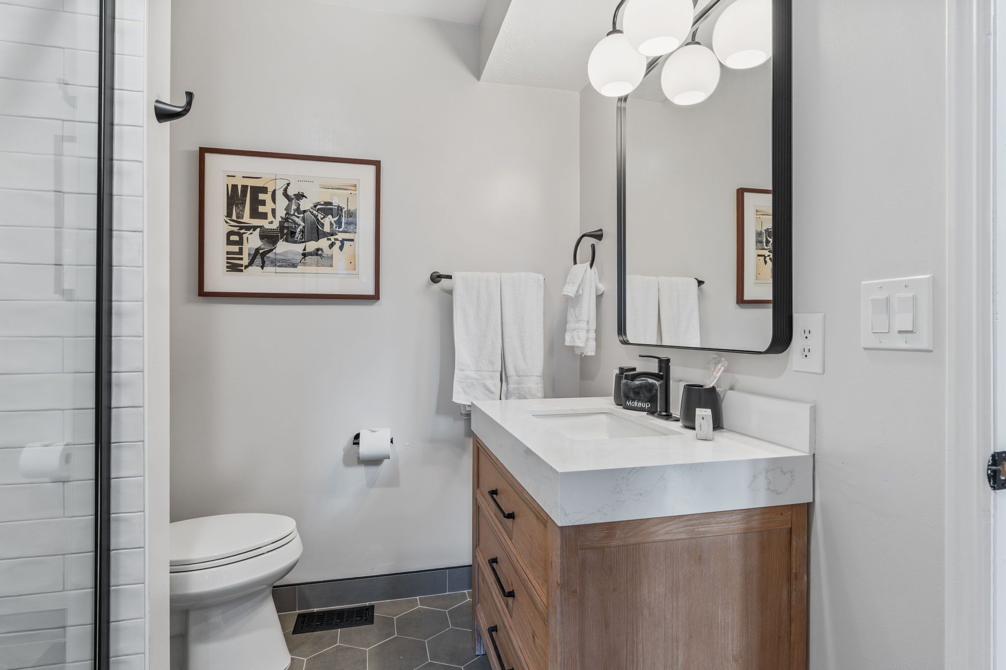 Un baño moderno con inodoro blanco, lavamanos de mármol, armarios de madera, un espejo grande y un cuadro en la pared.