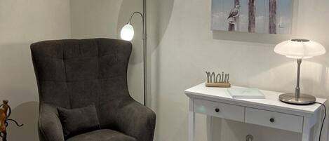 A modern living room with a dark grey upholstered armchair and ottoman, a white side table with a lamp, and a painting of seagulls on the wall.